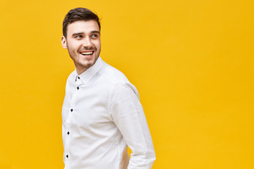 Wall Mural - Elegance, style and masculinity concept. Attractive trendy looking young brunette man with bristle and happy smile expressing confidence posing isolated at yellow studio wall, turning head round