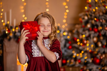 Wall Mural - little beautiful girl with white hair and in a red dress in the room dreams of gifts with her eyes closed. behind her is a Christmas tree in lights and a fireplace