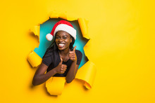 Young African Woman Wear In Santa Hat With Thumbs Up Standing In Yellow Torn Paper Hole Background