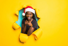 Young African Woman Wear In Santa Hat Pointed On You Standing In Yellow Torn Paper Hole Background