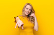 young pretty blonde woman smiling cheerfully and pointing to camera while making a call you later gesture, talking on phone against flat color wall