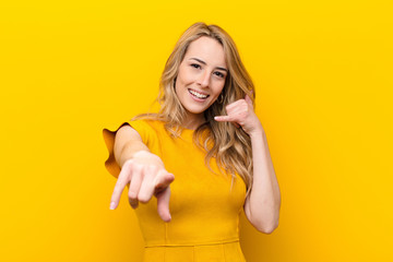 young pretty blonde woman smiling cheerfully and pointing to camera while making a call you later gesture, talking on phone against flat color wall