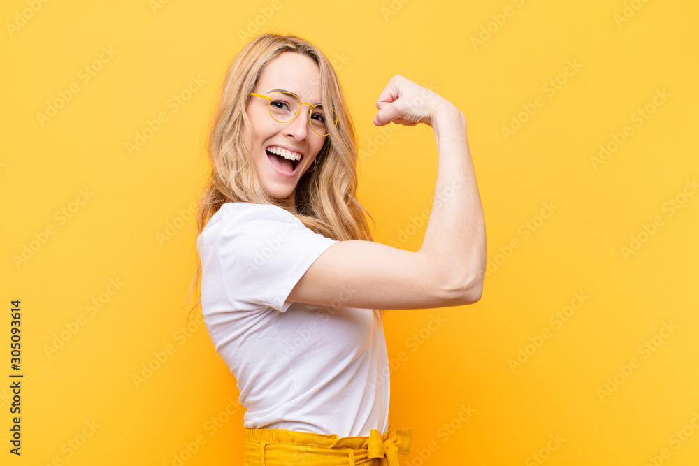 young pretty blonde woman feeling happy, satisfied and powerful, flexing fit and muscular biceps, looking strong after the gym against flat color wall - obrazy, fototapety, plakaty 