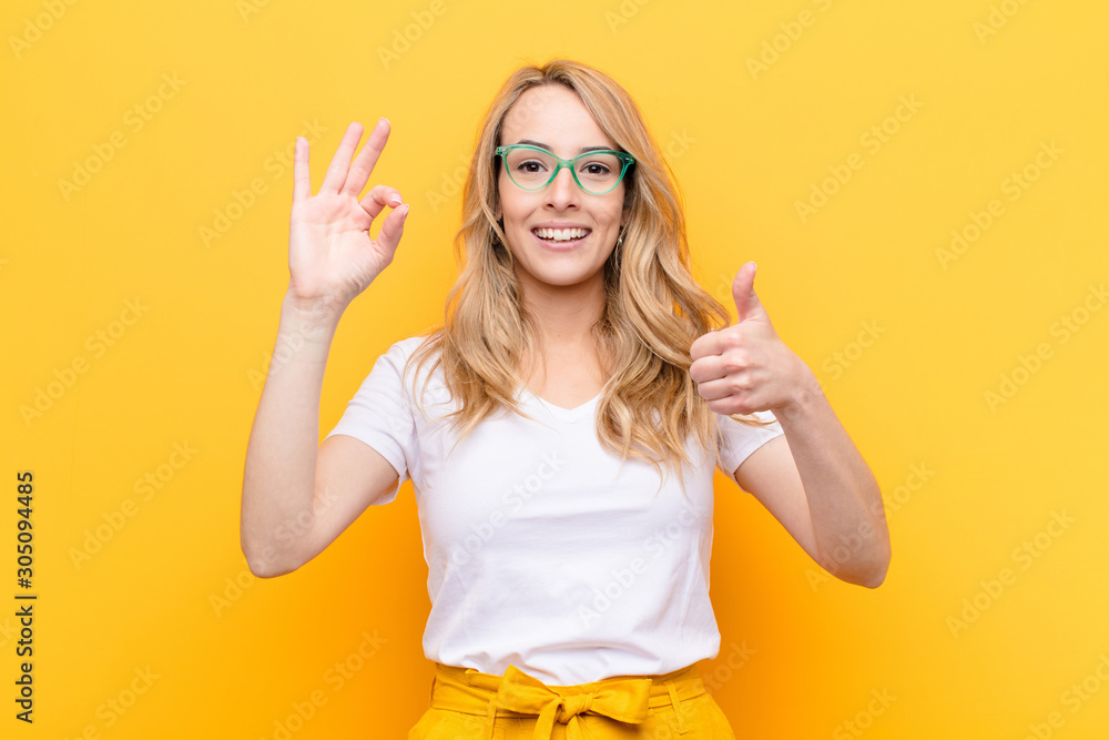 young pretty blonde woman feeling happy, amazed, satisfied and surprised, showing okay and thumbs up gestures, smiling against flat color wall - obrazy, fototapety, plakaty 