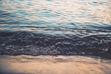 A small wave beats on the shore of the river, lake, sea. Yellow warm light from the setting sun. Close-up of the wave. Sand. Calm water. Background.