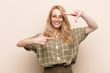 young pretty blonde woman feeling happy, friendly and positive, smiling and making a portrait or photo frame with hands against flat color wall