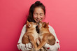 © wayhome.studio  - Joyful brunette young woman with pony tail holds two shiba inu puppies, expresses love to animals, wears sweater, stands over plane bright pink background. People, animals and friendship concept