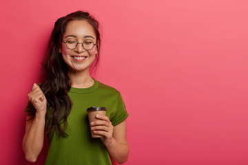 Wall Mural - Studio shot of joyful student raises fist, rejoices triumph and successfully finished task, has coffee break after long hours of work, smiles positively, wears eyeglasses, t shirt, poses with beverage