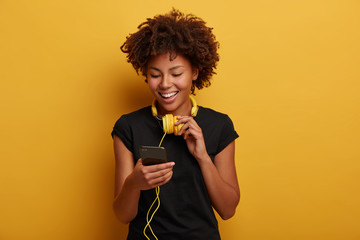 Wall Mural - cheerful curly girl picks up song in playlist, wears yellow headset around neck, black t shirt, smil