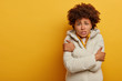 Dissatisfied dark skinned woman feels frozen after outdoor walk during frosty weather, wears warm white coat, trembles and keeps hands crossed over chest, stands against yellow background, copy space