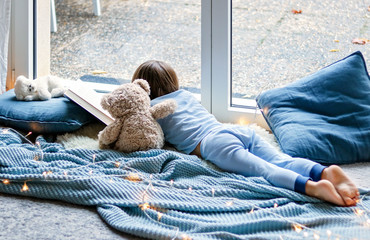 Cozy holidays at home. Cute little child lying on blue knitted blanket with teddy bear friend hugging him on floor at window reading book. Winter season lifestyle. Leisure time.