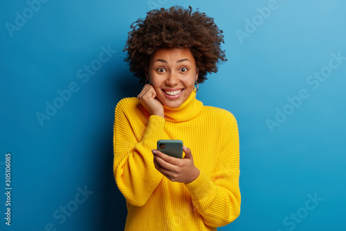 Joyous dark skinned young lady poses with mobile phone device, types text message on modern smartphone, watches funny video during free time, enjoys good internet connection, wears yellow sweater