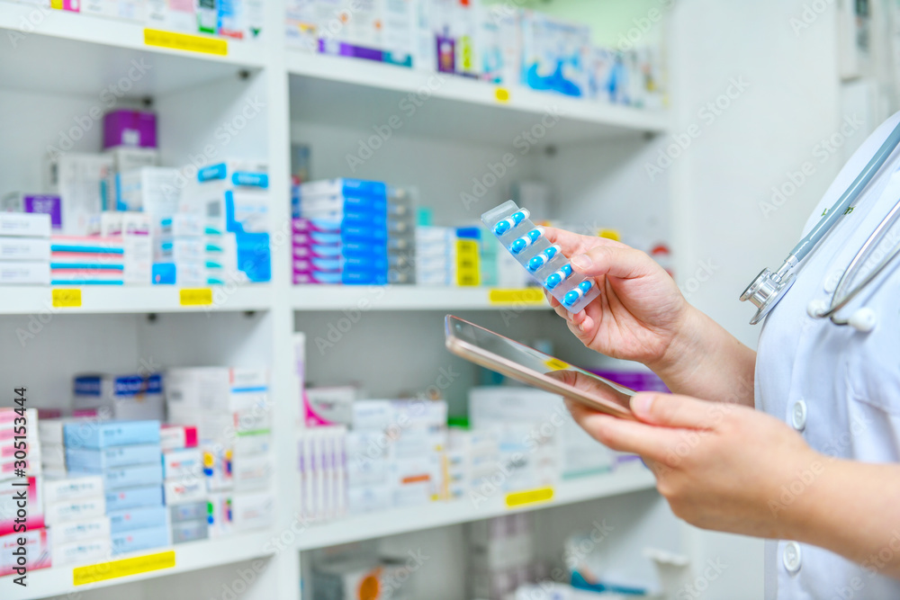 Doctor holding medicine capsule pack and computer tablet for filling prescription in pharmacy drugstore. - obrazy, fototapety, plakaty 