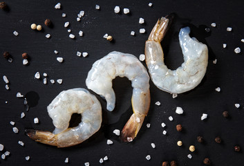 three large raw peeled shrimp with coarse sea salt on black background, top view