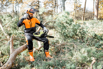 Poster - Portrait of a professional lumberman in protective workwear sitting with a chainsaw on the felled tree, resting after the hard work in the forest