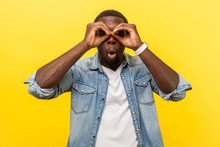 Portrait Of Astonished Young Man In Denim Casual Shirt Looking At Camera Through Fingers In Binoculars Gesture With Surprised Expression, Saying Wow. Indoor Studio Shot Isolated On Yellow Background