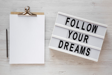 'Follow your dreams' words on a lightbox, clipboard with blank sheet of paper on a white wooden surface, top view. Overhead, from above, flat lay.