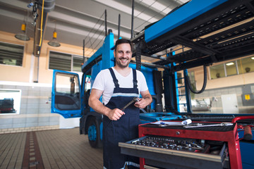 Portrait of positive smiling truck serviceman with tools standing by truck vehicle in workshop. Truck vehicle maintenance and servicing.