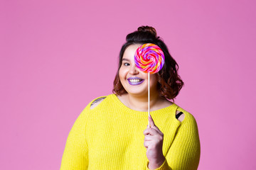 Cheerful plus size model with big lollipop, fat woman in yellow jumper on pink background