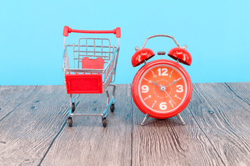 Shopping cart and classic alarm clock on blue background. Sale time buy mall market shop consumer concept. Selective focus.