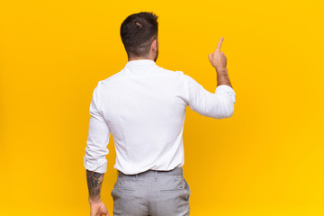 young handosme man standing and pointing to object on copy space, rear view against flat color wall