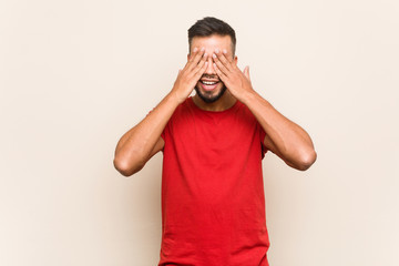 Wall Mural - Young south-asian man covers eyes with hands, smiles broadly waiting for a surprise.