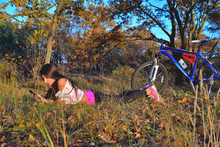 A Girl Lies On The Grass With A Phone In Her Hands, A Bicycle Is Standing Nearby In The Background, Autumn Sunny Evening, Forest Edge, Blue Sky