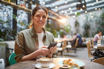 Portrait of beautiful young woman smiling at camera while using smartphone on outdoor terrace in cafe or coffee shop, copy space