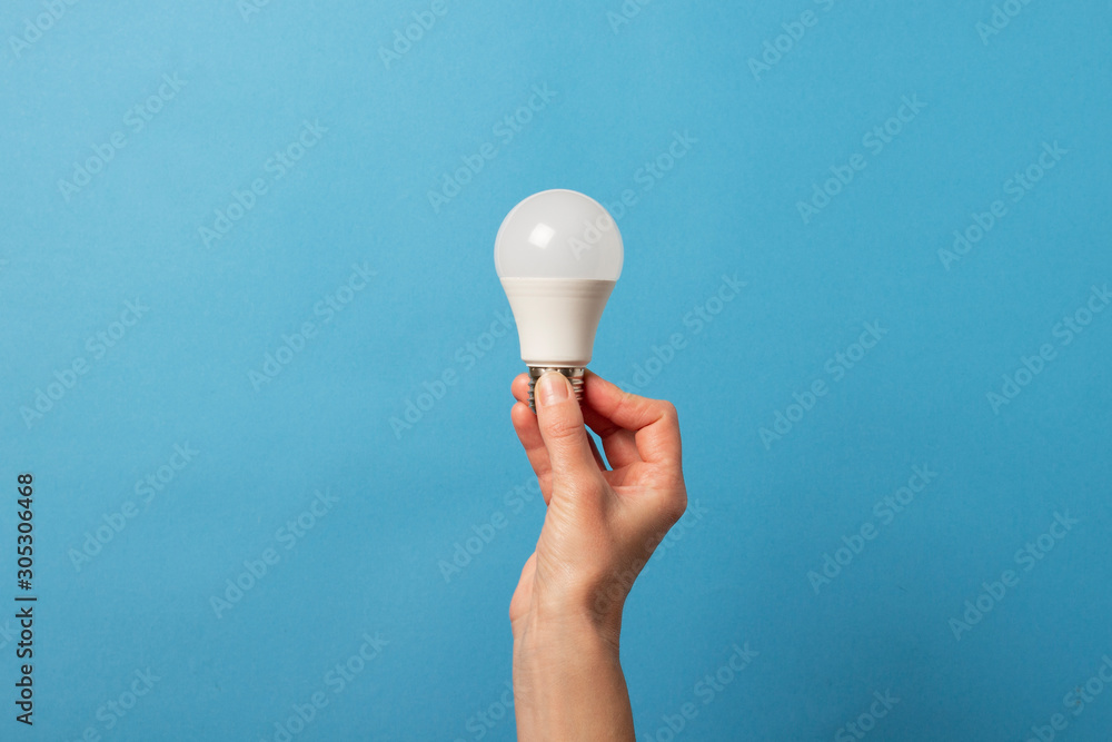 Female hand holds an LED lamp on a blue background. Energy saving concept, alternative energy sources, idea - obrazy, fototapety, plakaty 