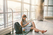 © Elizaveta - theme of tourism and travel of young student. Beautiful young caucasian girl in dress and hat sits on floor tourist rug inside terminal airport terminal. Waiting room delayed flight, delay departure