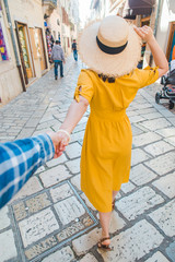 Wall Mural - follow me concept. woman in yellow sundress in straw hat walking forward by small resort city street holding man hand