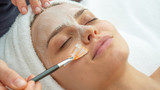 Fototapeta  - Close up of cosmetologist applies facial clay mask on an young beautiful woman face before ultrasonic cleaning procedure for skin pores and deep moisturizing.