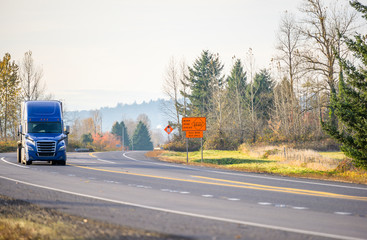 Big rig blue semi truck driving on the turning road with attention road work sign