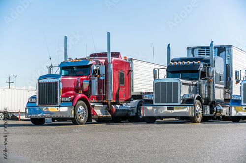 Classic bonnet big rigs semi trucks with reefer semi trailers standing in row on truck stop parking lot waiting for delivery schedule