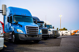 Different big rigs semi trucks standing in row on the truck stop parking lot at evening time