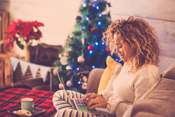 woman alone at home sitting on the sofa working or shopping with laptop and with cookies and tea or coffee and with christmas tree at the background