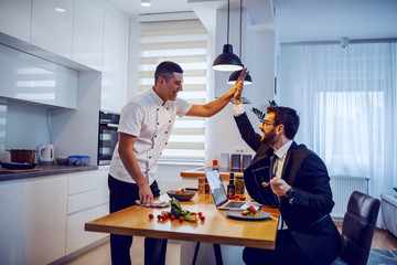 Bearded businessman in suit sitting at dining table, eating meal and giving high five to chef for perfect meal. Home interior.