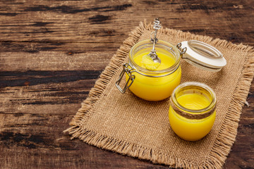 Pure or desi ghee (ghi), clarified melted butter. Healthy fats bulletproof diet concept or paleo style plan. Glass jar, silver spoon on vintage sackcloth. Wooden boards background