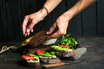 Wall Mural - Toast or sandwich with avocado, cheese, strawberries, herbs and seeds on a dark background. Female hands serve a dish. An idea for bruschetta or for a healthy snack. Healthy vegan breakfast.