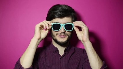 Canvas Print - Portrait of young smiling man puts on cyan sunglasses on pink background.