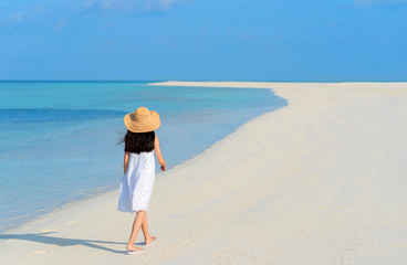 Back of young Asian girl walking on white sand beach with clear blue sea and sky. Teenager girl wearing sun straw hat, white dress enjoy vacation. Outdoor summer vacation travel concept, copy space.