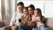 © fizkes - Happy parents with kids daughters relax on sofa using smartphone