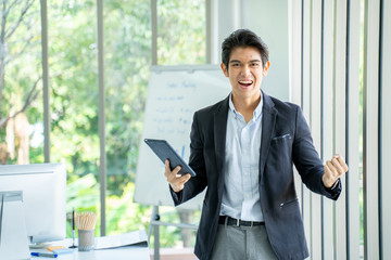 Excited young asian businessman happy and hurrah while standing in the office.Happy Asian man holding tablet and raising his arm up to celebrate success or achievement.