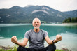 Leinwandbild Motiv A senior man pensioner sitting by lake in nature, doing yoga exercise.