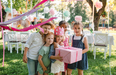 Portrait of small girl with friends and present outdoors in garden in summer.