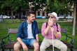 © Halfpoint - Young man and blind senior with white cane sitting on bench in park in city.