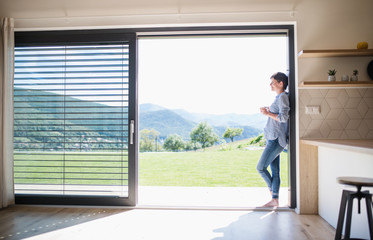 Side view of young woman with coffee standing by patio door at home.