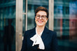 © Maskot - Portrait of confident smiling senior businesswoman against office