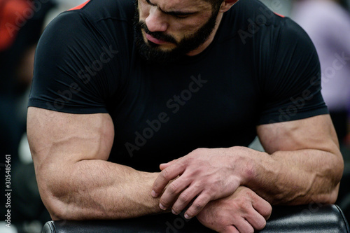 sport mental concentration concept of young strong mucle cute caucasian male with beard wearing black clothes leaning on simulator in athletic gym