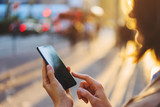 Fototapeta Perspektywa 3d - Young woman getting taxi via cab application on smartphone while standing on city street at evening. Woman texting message in chat on mobile phone walking outdoors. 4G internet